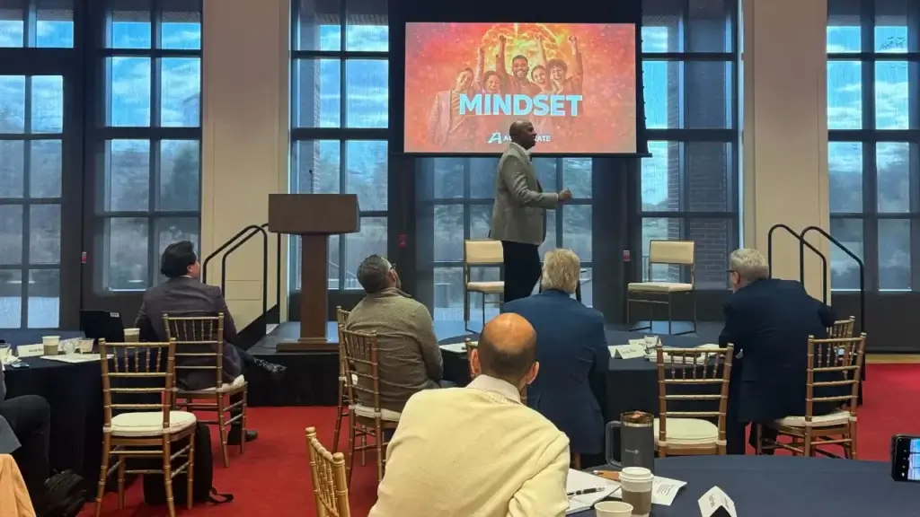 A sales leader presenting at a meeting with a "Mindset" slide on screen, with attendees seated at round tables