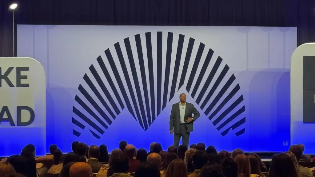 Walter Bond on stage at a keynote event, addressing a large seated audience with an arched striped backdrop behind him